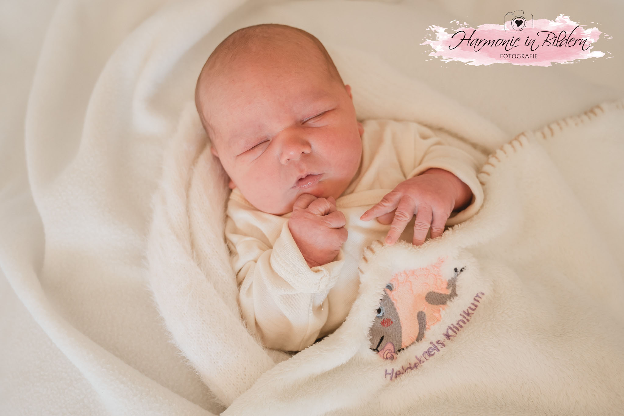 Ein Neugeborenes trägt ein weißes Strick-Oberteil und ist auf einer Decke mit Pflanzen-Motiven gebettet. Es schaut aus zugekniffenen Augen in Richtung Kamera, scheint zu lächeln und ballt die Hände zu Fäusten. Rechts oben im Bild ist eine Kennzeichnung in Form eines rosa Farbklexes zu sehen. Über dem Klex ist eine gezeichnete Kamera mit Herz im Objektiv abgebildet. Darunter steht handgeschrieben Harmonie in Bildern Fotografie,