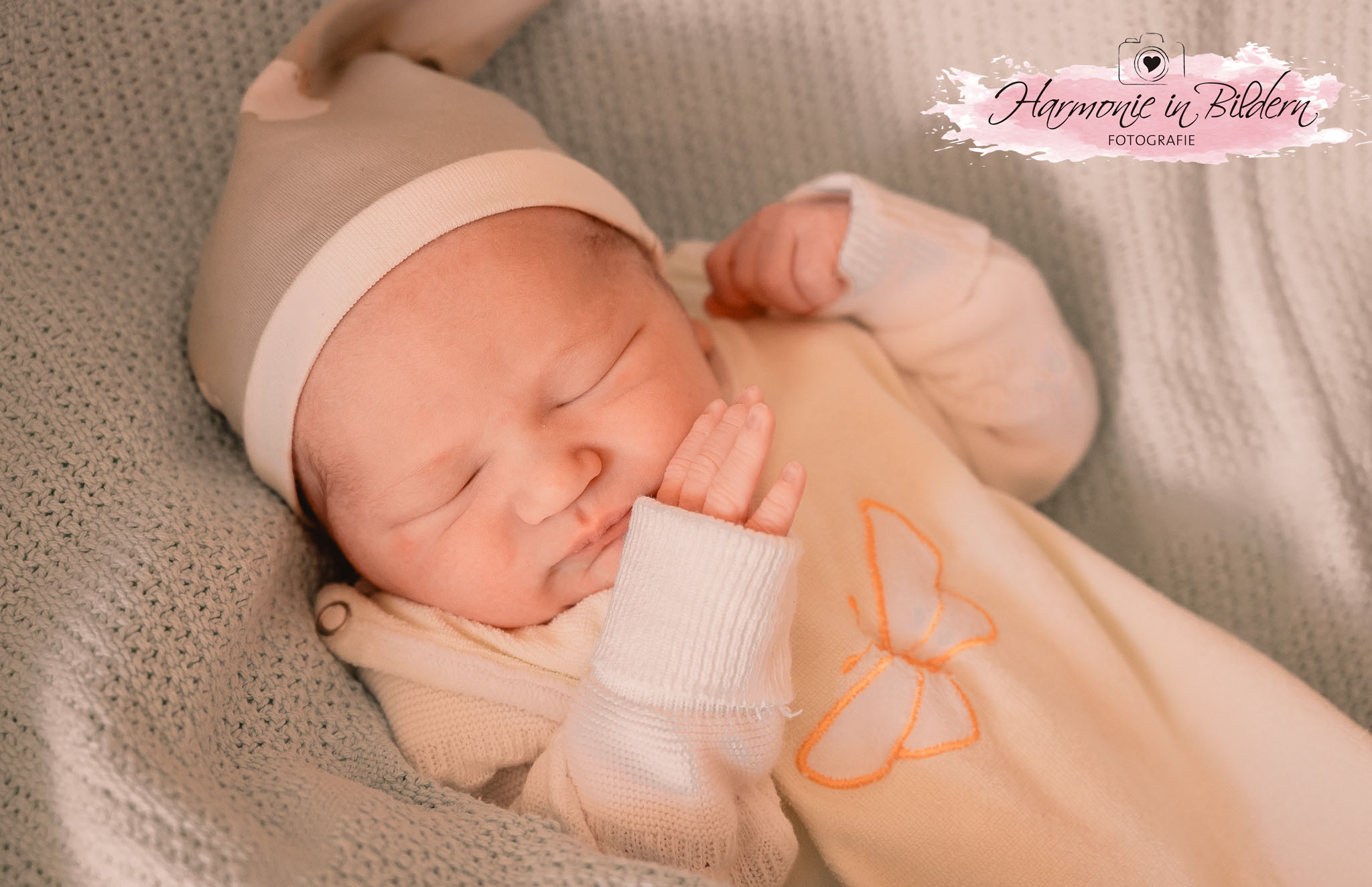 "Ein Neugeborenes mit beiger Mütze und orangenem Strampler mit Schmetterlingsmotiv schläft auf einer grauen Strickdecke. Rechts oben im Bild ist eine Kennzeichnung in Form eines rosa Farbklexes zu sehen. Über dem Klex ist eine gezeichnete Kamera mit Herz im Objektiv abgebildet. Darunter steht handgeschrieben Harmonie in Bildern Fotografie "