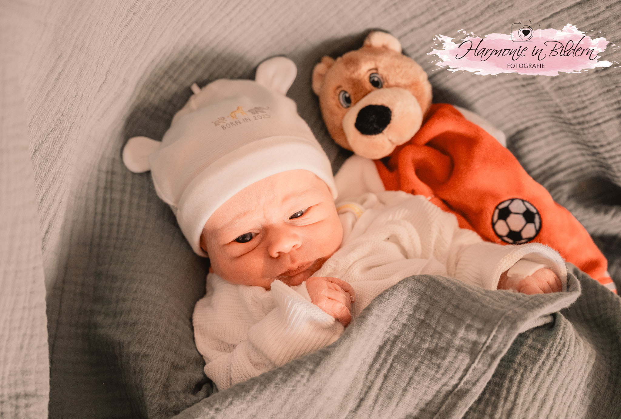 Das Bild zeigt ein Neugeborenes auf einer groben grauen Decke. Es hat ein weißes Oberteil und eine weiße Mütze mit Bärchen-Ohren an, auf der ein Schriftzug zu sehen ist: Born in 2025. Das Kind schaut mit verquollenen Augen nach unten. Es hat ein Pflaster auf der linken Hand. Links neben dem Kind liegt ein Plüsch-Teddy mit orangenem Körper und Fußballmotiv. Rechts oben im Bild ist eine Kennzeichnung in Form eines rosa Farbklexes zu sehen. Über dem Klex ist eine gezeichnete Kamera mit Herz im Objektiv abgebildet. Darunter steht handgeschrieben Harmonie in Bildern Fotografie
