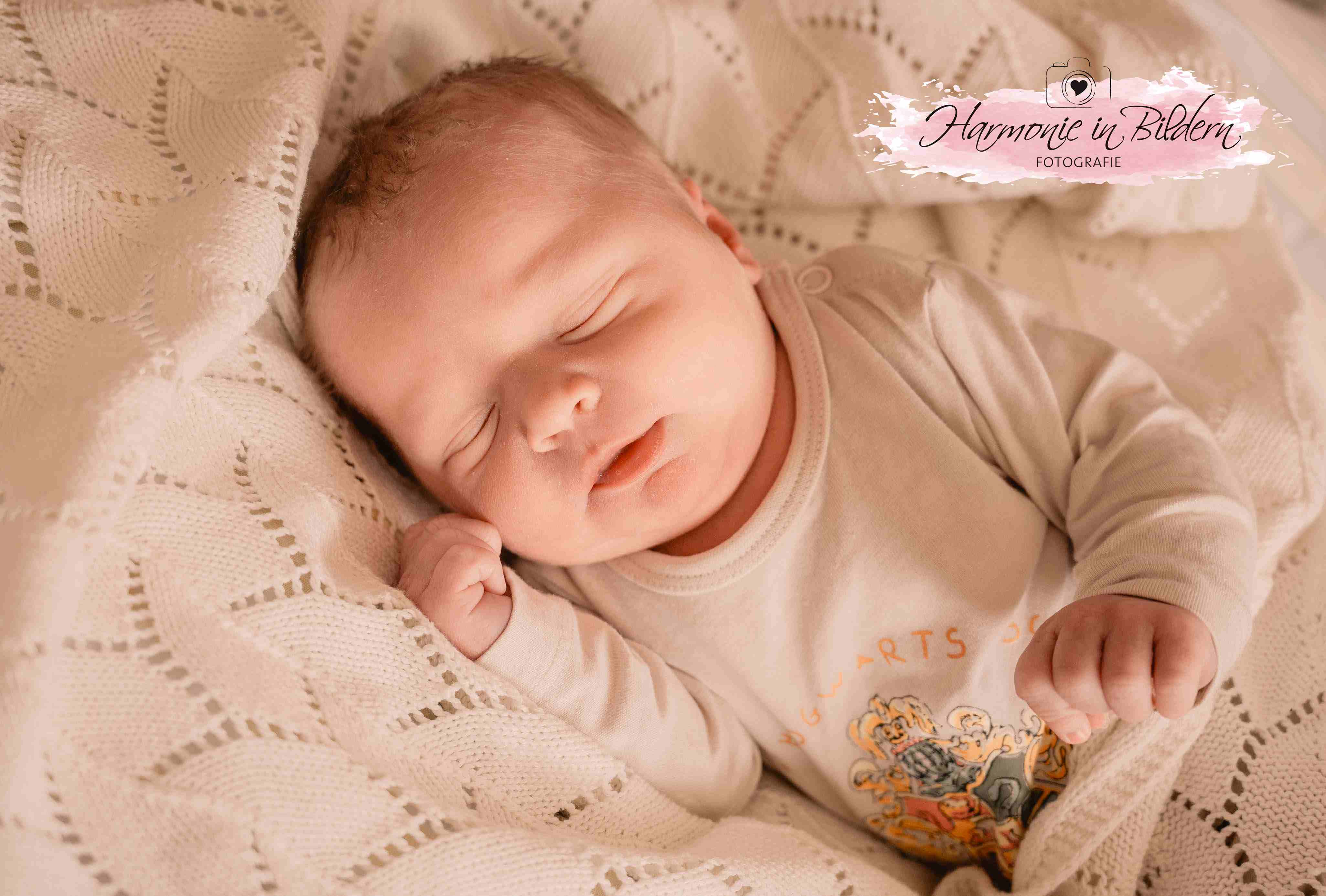 Ein Neugeborenes schläft friedlich auf einer weißen Strickdecke. Rechts oben im Bild ist eine Kennzeichnung in Form eines rosa Farbklexes zu sehen. Über dem Klex ist eine gezeichnete Kamera mit Herz im Objektiv abgebildet. Darunter steht handgeschrieben Harmonie in Bildern Fotografie